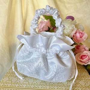 May include: A white satin drawstring bag with a ruffled top and a gathered handle. The bag features an embroidered floral design and is tied with white ribbons. Pink and white roses are visible inside the bag.