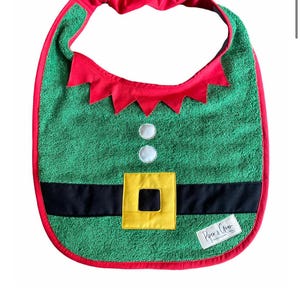 Könnte beinhalten: Grünes Lätzchen im Elfen-Design mit roter Einfassung und gelber Schnalle. Das Lätzchen hat einen roten Kragen mit gezackten Kanten und zwei weißen Knöpfen. Der Text "I AM THE CUTEST ELF CHRISTMAS BIB" steht unten.
