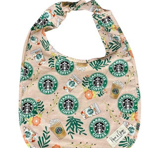 Könnte beinhalten: Ein rosa Stoff-Lätzchen für Babys mit einem sich wiederholenden Muster aus Starbucks-Kaffeebechern, grünen Blättern und orangefarbenen Blumen. Das Lätzchen hat einen grünen Saum und ein weißes Etikett mit der Aufschrift "Piper & Gray".