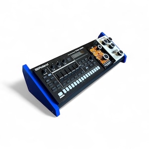 Puede incluir: Dispositivo negro de producción musical electrónica con un soporte de madera azul. El dispositivo tiene numerosas perillas, botones y una pantalla. El logotipo de Roland es visible. Módulos adicionales con perillas y un gráfico de tigre.