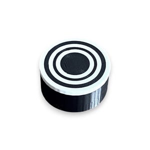May include: A circular object with a black and white target design. The top has concentric circles in black and white. The sides are black. The object is set against a white background.