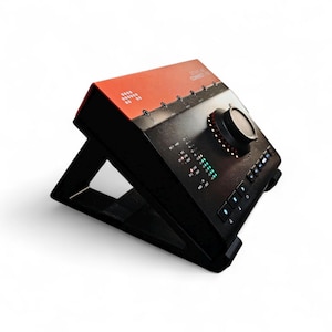 May include: A black and orange audio interface with a large control knob and multiple buttons. The device is angled on a black stand. The top of the interface has the text "Apollo Twin Connect".