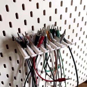 Puede incluir: Panel perforado blanco con múltiples cables de audio de varios colores y tamaños. Los cables se sujetan con soportes de plástico blanco. El panel perforado tiene agujeros espaciados uniformemente.