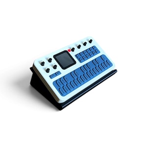 Puede incluir: Un controlador de sintetizador de música electrónica blanco y azul con una pantalla cuadrada central, cuatro perillas giratorias negras y una cuadrícula de botones rectangulares azules sobre un soporte negro inclinado.