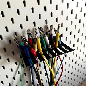 May include: A selection of multi-coloured audio cables with metal connectors, arranged in black holders against a white pegboard. Cables are in various colours, including red, yellow, blue, and green.