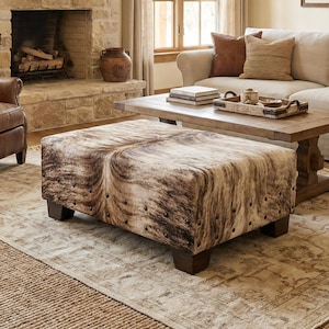 May include: A living room with a rustic theme. Features include a stone fireplace, a cowhide ottoman, leather chairs, a beige sofa, and a wooden coffee table. Decorative items include a steer skull and framed artwork.