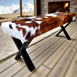 May include: A bench with a cowhide seat in shades of brown and white, supported by black, X-shaped legs. The bench is placed in front of a window with a mountain view and a fireplace.
