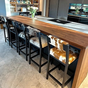 May include: A long wooden kitchen island with a black countertop and a row of black bar stools with cowhide seats. The stools are arranged in a row along the island, facing the kitchen.