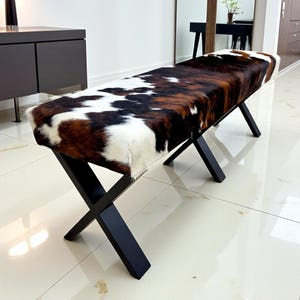 May include: A bench with a cowhide seat in shades of brown, black, and white, atop black, crossed legs. The bench is a decorative furniture piece, suitable for a living room or entryway.