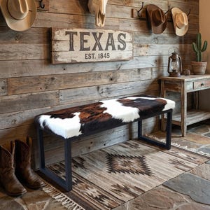 Puede incluir: Entrada rústica con un banco de piel de vaca, un letrero de Texas y un cráneo de longhorn. Sombreros de vaquero cuelgan de la pared de madera. Una alfombra estampada y botas de vaquero completan la decoración.