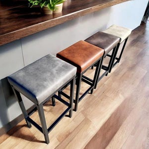 May include: Four bar stools with black metal frames and upholstered seats in different colours: grey, brown, tan, and beige. The stools are arranged in a row on a wooden floor.