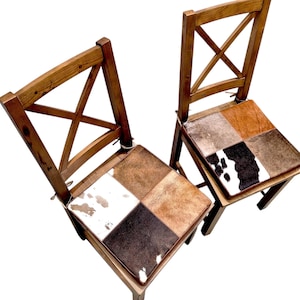 Könnte beinhalten: Zwei Holzstühle mit einem Patchwork-Sitzkissen aus Kuhfell. Die Stühle haben ein schlichtes, rustikales Design mit einem Kreuzmuster auf der Rückenlehne.