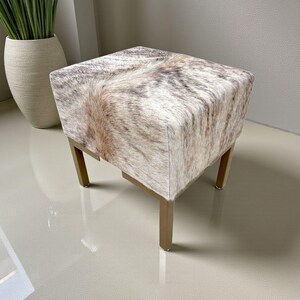May include: A square ottoman with a cowhide pattern in shades of brown, tan, and white. The ottoman has a wooden frame with four legs. The design is a mix of natural and neutral tones, suitable for various interior styles.