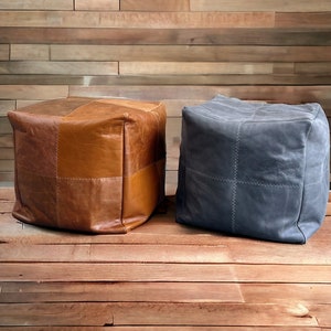 May include: Two square leather poufs, one brown and one grey, with a stitched pattern. The poufs are sitting on a wooden floor.