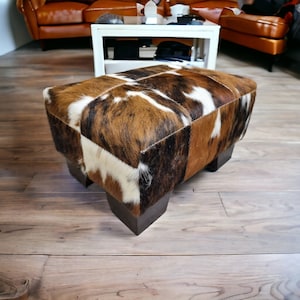 May include: A brown, white, and black cowhide ottoman with a square top and four legs. The ottoman is sitting on a wooden floor.