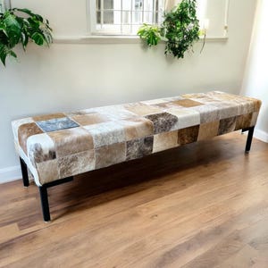 May include: A long bench with a patchwork design made of various shades of brown, tan, and cream-colored leather squares. The bench has black metal legs and is placed on a hardwood floor. The bench is approximately 60 inches long.