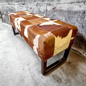 May include: A brown and white cowhide bench with a dark brown wooden frame. The bench is made of patchwork pieces of cowhide.