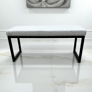 May include: A modern bench with a rectangular, light gray, textured seat. The seat is supported by a black, angular metal frame. The bench is placed in a room with white walls and a marble floor.