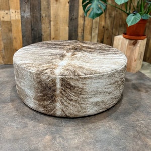 May include: A round, brown and white cowhide ottoman with a soft, plush texture. The ottoman is sitting on a brown floor.