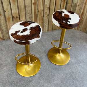 May include: Two gold bar stools with brown and white cowhide seats. The stools have a simple design with a round seat and a single, vertical support.