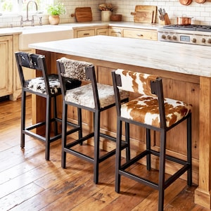 Könnte beinhalten: Drei Barhocker mit schwarzen Metallrahmen und Kuhfell-Sitzen in einer Küche. Die Kuhfellmuster variieren, mit schwarz-weißen, grau-weißen und braun-weißen Designs. Die Hocker stehen an einer Kücheninsel.