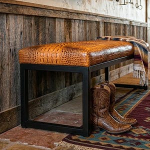 Op de afbeelding: Een bank met een bruine, getextureerde, imitatielederen bovenkant en een zwart metalen frame. Een paar bruine cowboylaarzen staat onder de bank. Een kleurrijke deken ligt op de bank.