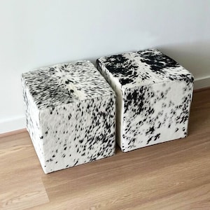 Puede incluir: Dos otomanas cúbicas de piel de vaca en blanco y negro. Las otomanas están hechas de un material suave y mullido y tienen un patrón único y natural.