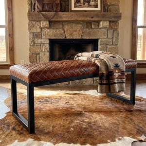 May include: A brown leather bench with a diamond-stitched pattern sits on a black metal frame. A Southwestern-style blanket rests on the bench. The bench is placed on a cowhide rug in front of a stone fireplace.