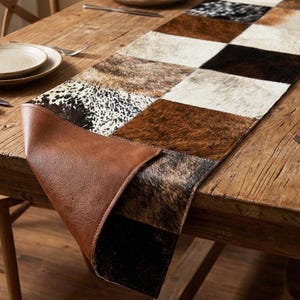 May include: A patchwork table runner with a cowhide design. The runner features squares of brown, black, and white cowhide, with a brown leather backing. The runner is placed on a wooden table, with plates and silverware visible.