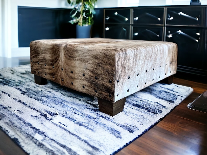 May include: A brown and white cowhide ottoman with metal studs sits on a blue and white striped rug.