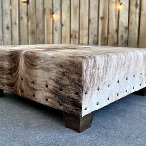 May include: A square, brown and white cowhide ottoman with dark brown wooden legs. The ottoman is decorated with metal studs around the edges.