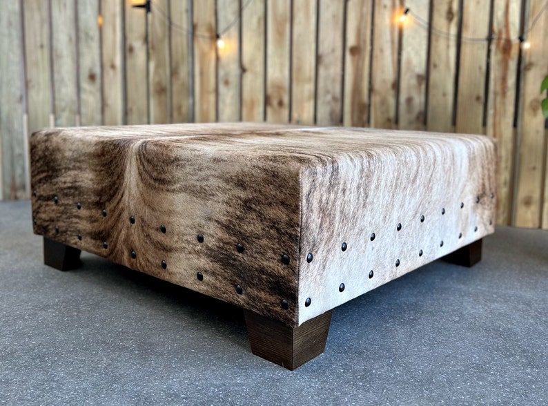 May include: A square, brown and white cowhide ottoman with dark brown wooden legs. The ottoman is decorated with metal studs around the edges.