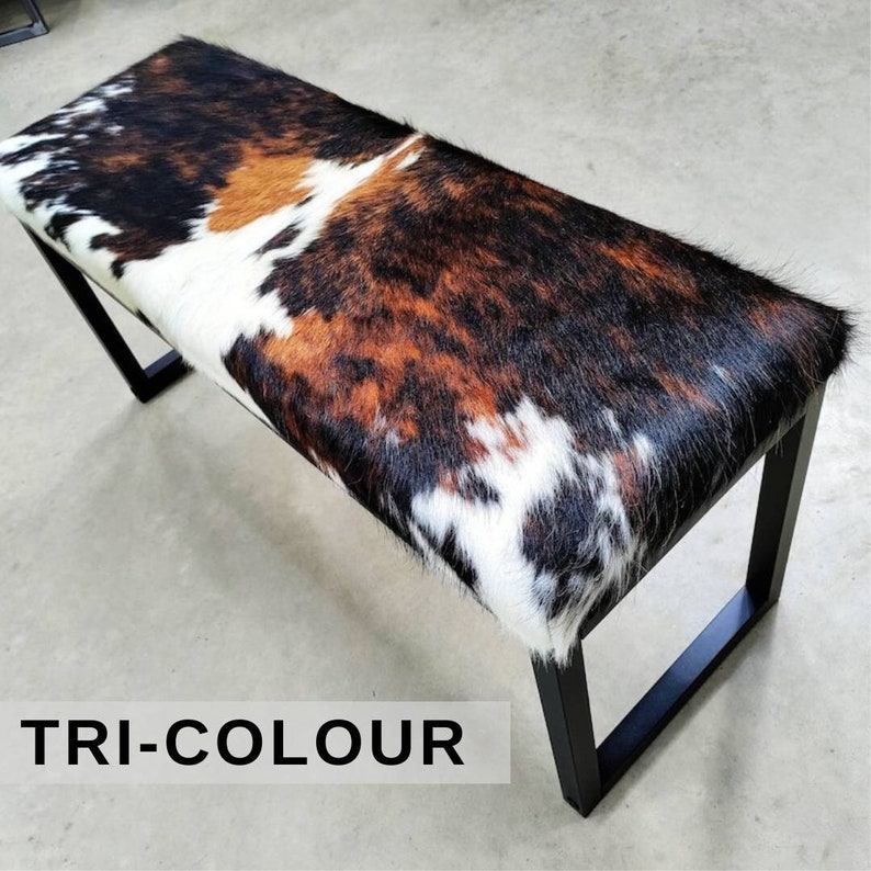 May include: A black metal bench with a tri-colour cowhide seat. The seat is white, brown, and black. The bench has a simple, modern design.
