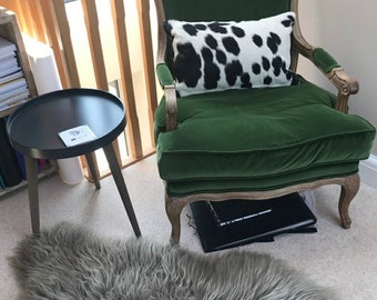 Puede incluir: Un sillón de terciopelo verde con un cojín estampado de piel de vaca en blanco y negro. El sillón tiene patas de madera y está sobre una alfombra gris claro. Una mesa auxiliar negra con tapa redonda está en el fondo.