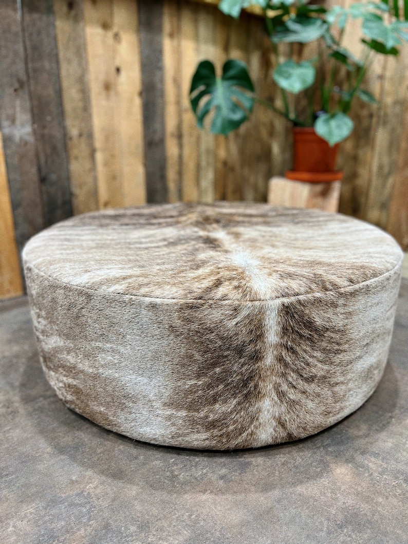May include: A round, brown and white cowhide ottoman with a soft, fuzzy texture.