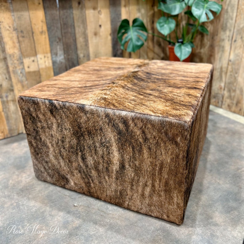 Pu&ograve; includere: Un pouf quadrato rivestito in pelle di mucca marrone e nera con un leggero bagliore. Il pouf &egrave; appoggiato su un pavimento di cemento.