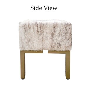 May include: Side view of a square ottoman with a textured, light-colored fur-like top. The ottoman has a gold-colored metal base with four legs. The text "Side View" is at the top of the image.