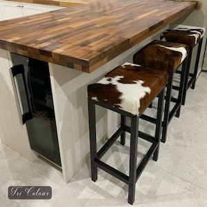 Peut inclure: Trois tabourets de bar en m&eacute;tal noir avec des si&egrave;ges en peau de vache brune et blanche. Les tabourets sont dispos&eacute;s en rang&eacute;e devant un comptoir en bois.