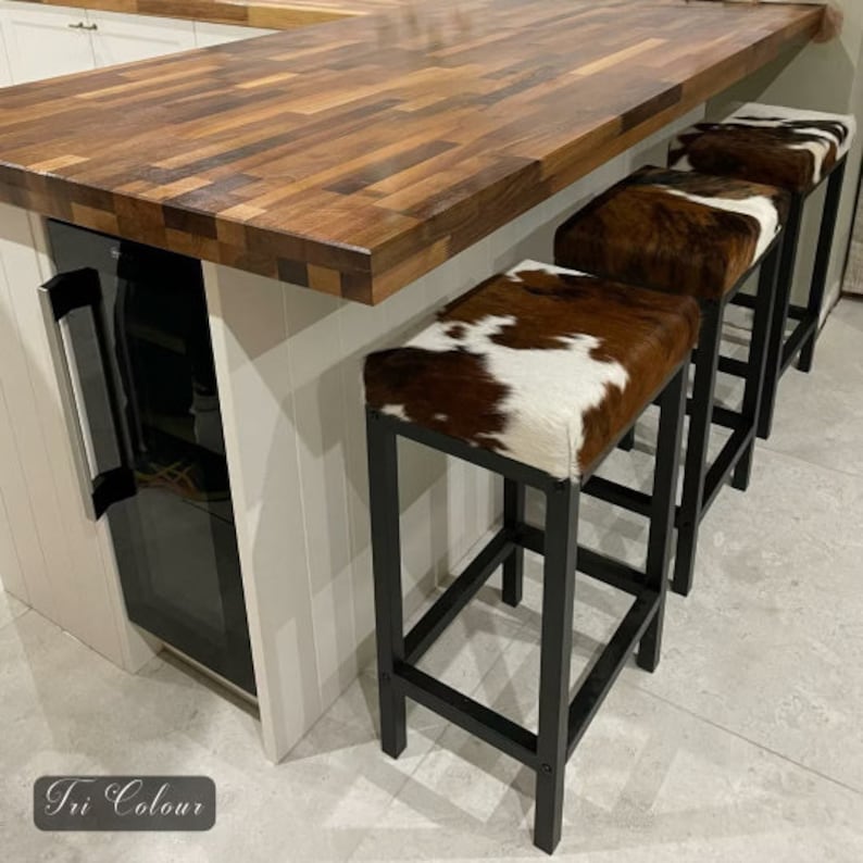 Peut inclure: Trois tabourets de bar en m&eacute;tal noir avec des si&egrave;ges en peau de vache brune et blanche. Les tabourets sont dispos&eacute;s en rang&eacute;e devant un comptoir en bois.