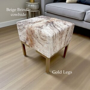 May include: A square ottoman with a beige brindle cowhide top and gold-colored legs. The cowhide has a natural pattern of brown and cream colors. The legs are a simple, modern design.