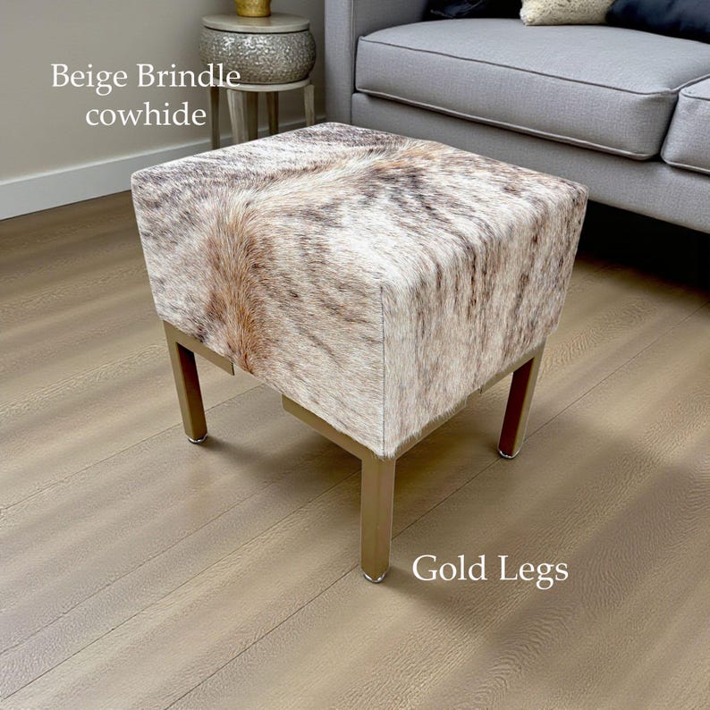 May include: A square ottoman with a beige brindle cowhide top and gold-colored legs. The cowhide has a natural pattern of brown and cream colors. The legs are a simple, modern design.