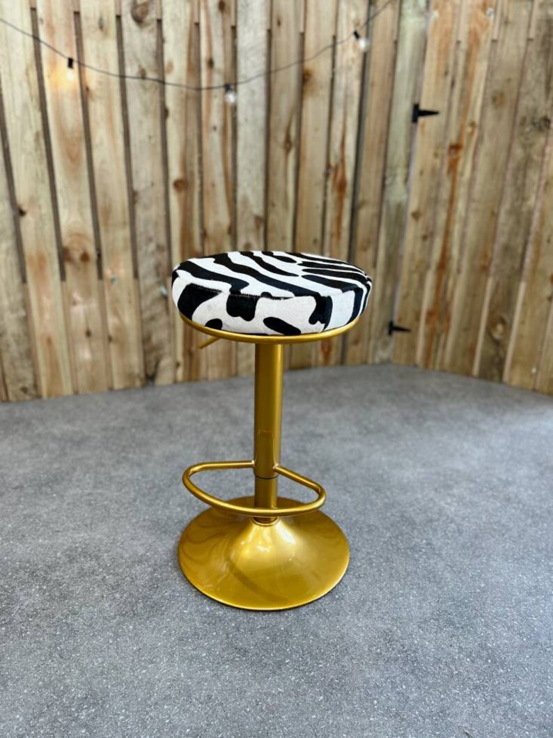 Op de afbeelding: Een gouden barstoel met een zebraprint zitkussen. De stoel heeft een ronde basis en een voetensteun.