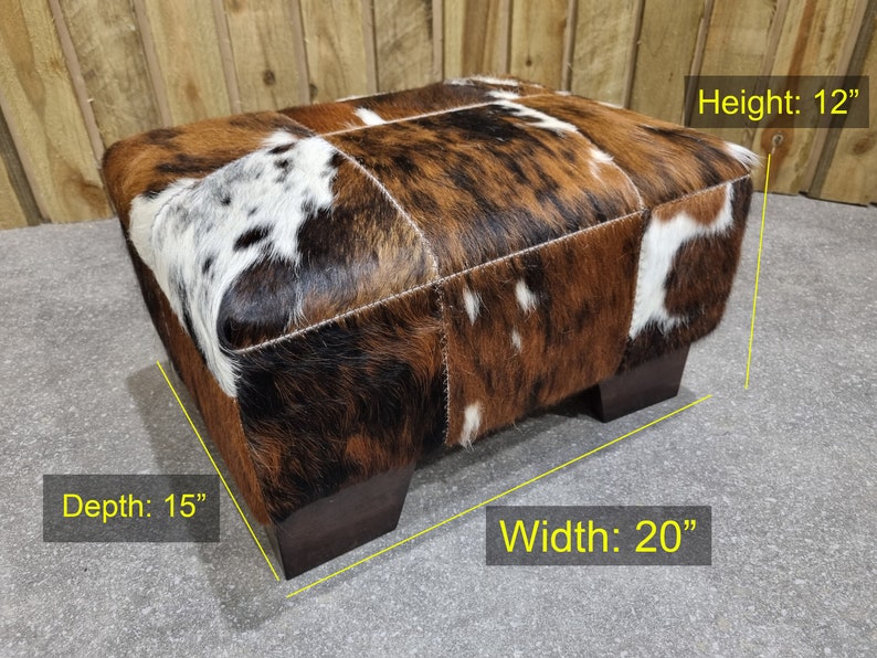 May include: A brown and white cowhide ottoman with a square top and four wooden legs. The ottoman measures 20 inches wide, 15 inches deep, and 12 inches high.