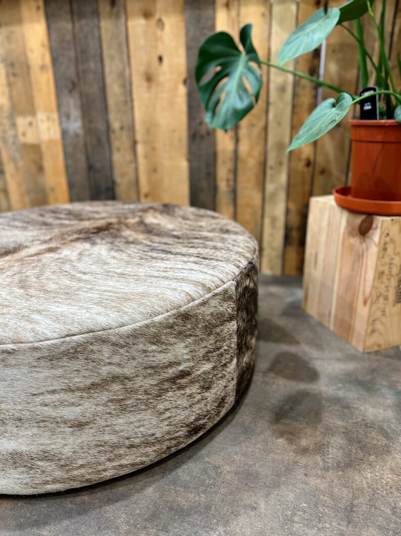 May include: A round, brown and white cowhide ottoman with a wooden background and a potted plant.