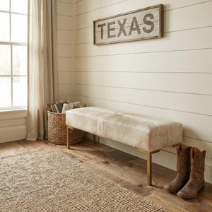 May include: A rectangular bench with a light-colored, textured hide upholstery and four slim, metallic gold-toned legs. The bench is positioned near a woven basket and a pair of brown leather cowboy boots on a wooden floor.