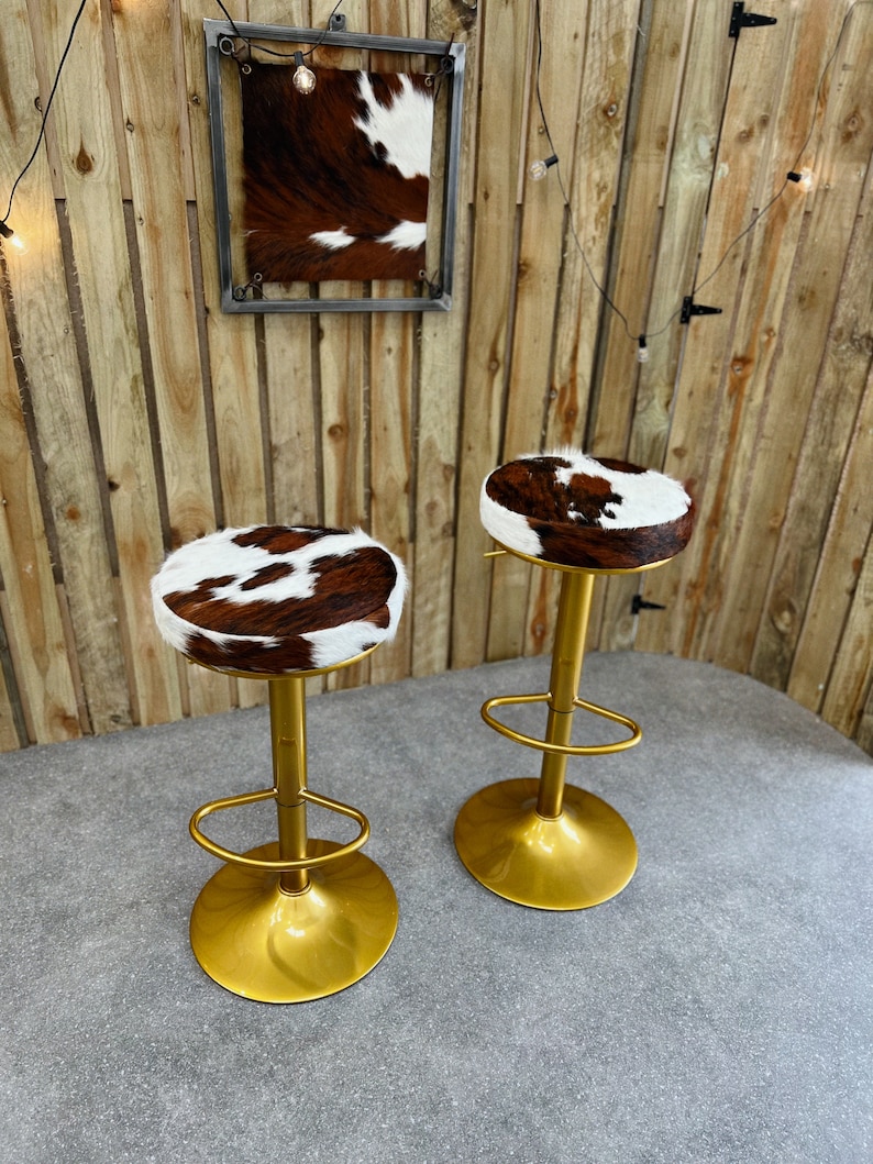 K&ouml;nnte beinhalten: Zwei goldene Barhocker mit braunen und wei&szlig;en Kuhfell-Sitzen. Die Hocker haben eine runde Basis und eine Fu&szlig;st&uuml;tze.