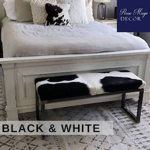 May include: Black and white cowhide bench with a black metal frame. The bench is sitting in front of a white bed with a white and black cowhide pillow. The text "Rose Maye DECOR" is visible in the upper right corner of the image.