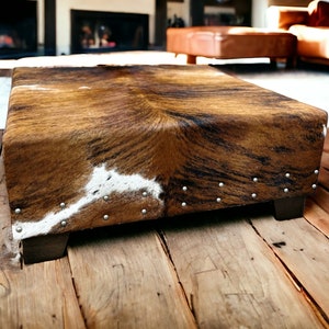 May include: A brown and white cowhide ottoman with silver studs. The ottoman is sitting on a wooden floor.