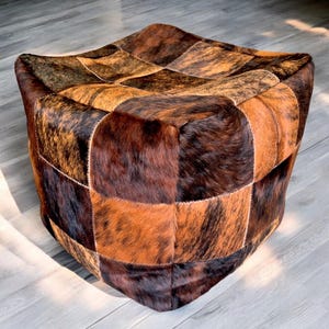 May include: A square-shaped ottoman with a patchwork design made from different shades of brown cowhide. The ottoman is made of leather and has a soft, plush texture.