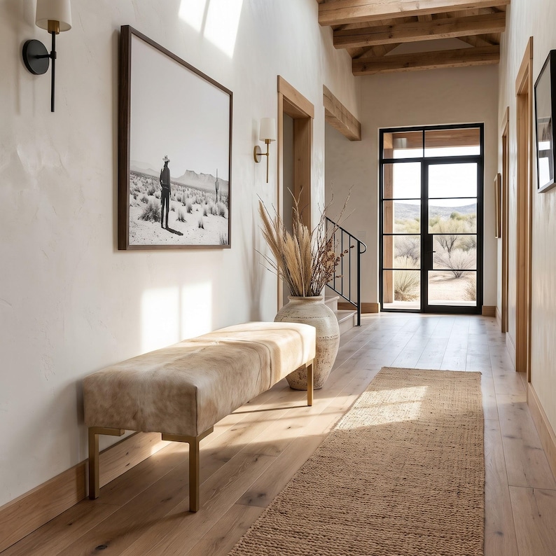 May include: A rectangular bench with a light tan, mottled hide-style upholstered seat and a simple gold-toned metal frame. The bench is positioned in a hallway with light-colored walls, a wooden floor, and a large framed black and white desert print.